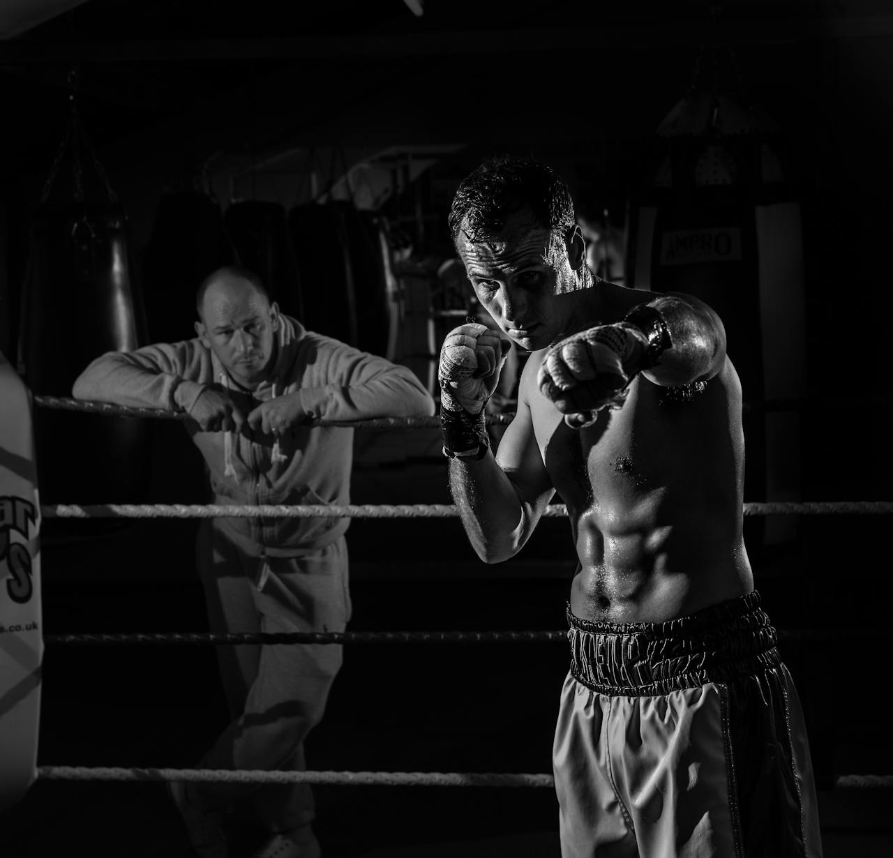 Boxer practicing in the ring with coach observing, captured in dramatic lighting.